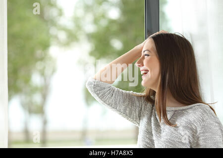 Seitliche Sicht auf ein fröhliches Mädchen Lachen mit Hand am Kopf und im freien Blick durch ein Fenster zu Hause oder im Hotelzimmer Stockfoto