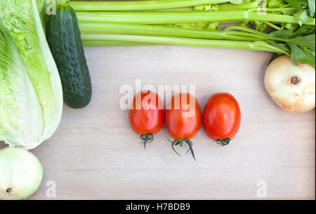 Drei Tomaten umgeben von frischem Gemüse (Zwiebel, Gurke, Salat und Sellerie Blätter) auf Holzbrett Stockfoto