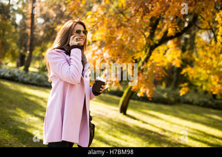 Junge Frau in den rosa Mantel im Gespräch über das Smartphone und hält Kaffee in der hand im Herbst Park gehen Stockfoto