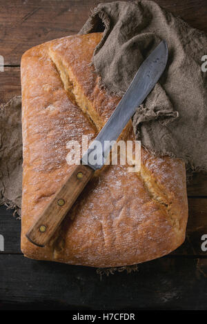 Ganze frisch gebackenes Handwerker Brot unter Sackleinen mit Vintage Messer über alten hölzernen Hintergrund. Draufsicht. Stockfoto
