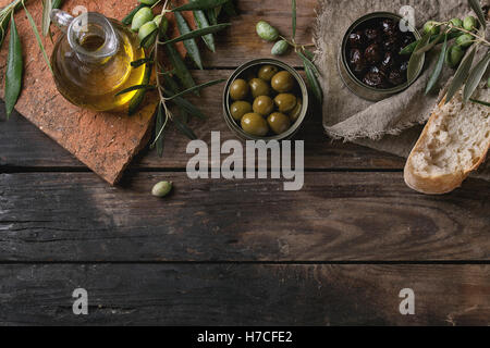 Grüne und schwarze Oliven in Blechdosen mit frischem Brot und junge Oliven verzweigen auf Sackleinen, Flasche Olivenöl auf Ton Bo Stockfoto