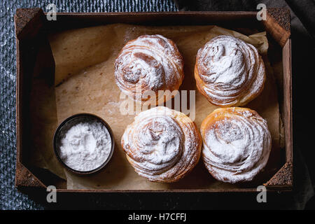 Moderne Gebäck Cruffins, ganzen und in Scheiben schneiden, wie Croissants und Muffins mit Puderzucker, serviert in Holzkiste auf schwarzem Textil-nap Stockfoto