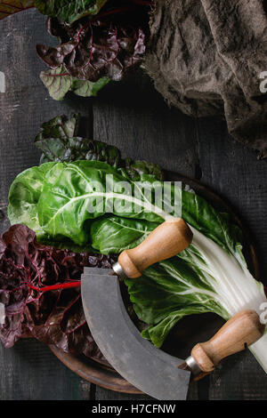 Vielzahl von frischen Mangold Mangold Salat Blätter auf Woode Schneidebrett mit Vintage Messer und Sackleinen Lappen über alten dunklen hölzernen ba Stockfoto