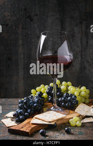 Die Trauben reif nasse rote und weiße Trauben mit Cracker Snack und Glas Rotwein auf Oliven Holz Schneidebrett über alte dunkle woo Stockfoto