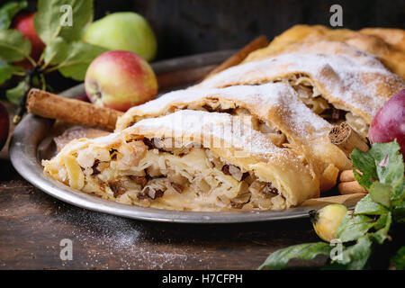 Hausgemachter Apfelstrudel, serviert mit frischen Äpfeln mit Blättern geschnitten, Metall-Zimtstangen und Puderzucker auf Vintage Tablett über Stockfoto