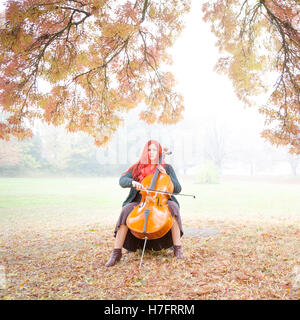 Porträt einer rothaarige Frau posiert mit einem Cello unter Herbst Bäume und braun/Orange Laub im West London Park, UK. Stockfoto