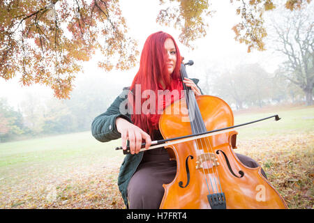 Porträt einer rothaarige Frau posiert mit einem Cello unter Herbst Bäume und braun/Orange Laub im West London Park, UK. Stockfoto