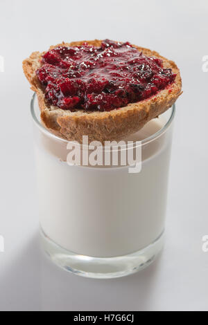 Geröstetes Brot mit Schicht aus schwarzen Johannisbeeren Marmelade an der Spitze der farblosen Glas voll von Kuhmilch auf weißem Hintergrund Stockfoto