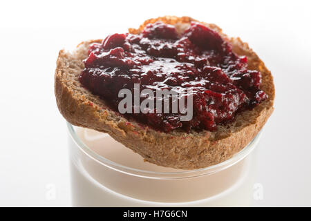 Geröstetes Brot mit Schicht aus schwarzen Johannisbeeren Marmelade an der Spitze der farblosen Glas voll von Kuhmilch auf weißem Hintergrund Stockfoto