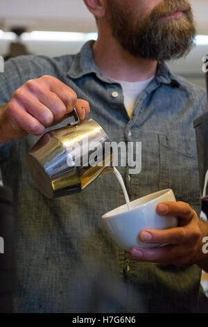 Barista mit einem Bart gießt Latte mit Schaum Milch in eine Tasse in einem Café. Stockfoto