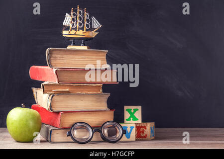 Alte Schulbücher am rustikalen Holztisch vor dem Hintergrund der schwarzen Tafel. Professoralen Gläser und Würfel mit Buchstaben. Stockfoto