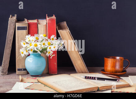 Das offene alte Buch liegend auf dem Holztisch unter Vintage Umschläge, Vase mit Gänseblümchen und eine Tasse mit einem heißen Getränk. Rustikale noch Stockfoto
