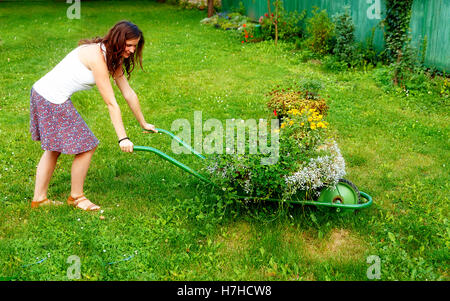 junge Frau im Garten spielerisch drängen dekorative Schubkarre voller Blumen. Stockfoto