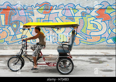 Havanna, Kuba - 18. Mai 2011: Junge kubanische Mann fährt eine Velo-Rikscha hinter einer Wand von Graffiti auf einer Straße in Zentral-Havanna. Stockfoto