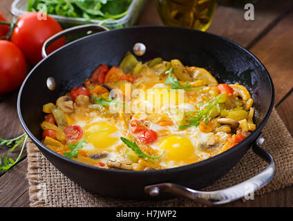 Spiegeleier mit Gemüse in einer Pfanne auf einem hölzernen Hintergrund. Stockfoto