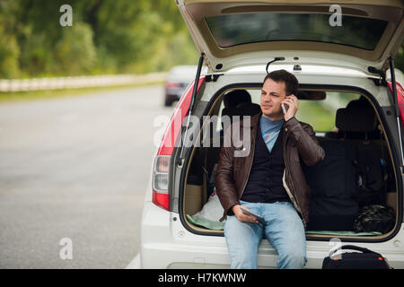 Freunde anzurufen. Glücklich Jüngling Handy sprechen und sitzt auf einem Kofferraum eines Autos Stockfoto