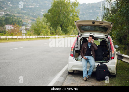 Freunde anzurufen. Glücklich Jüngling Handy sprechen und sitzt auf einem Kofferraum eines Autos Stockfoto