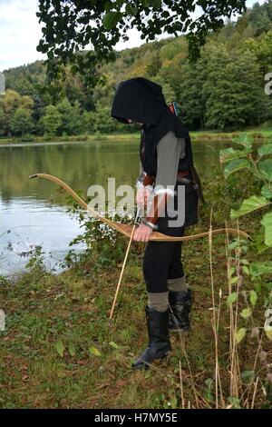 Mittelalterlicher Bogenschütze mit schwarzer Kapuze mit gespannten Bogen und Pfeile im See steht und schaut nach unten Stockfoto