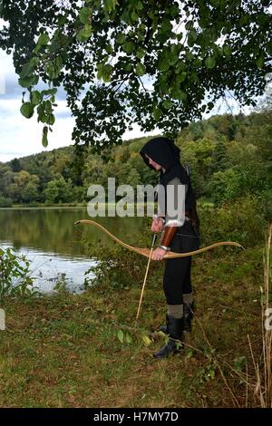 Mittelalterlicher Bogenschütze mit schwarzer Kapuze steht mit gespannten Bogen und Pfeile im See Stockfoto