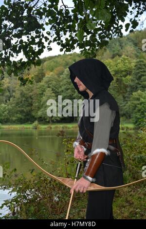 Mittelalterlicher Bogenschütze mit schwarzer Kapuze steht mit gespannten Bogen und Pfeile im See Stockfoto