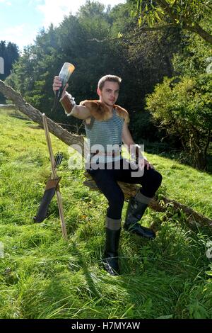 Mittelalterliche junge Mann, gekleidet hält Trinkhorn in der Hand und sitzt hoch auf Zweig in der Natur Stockfoto