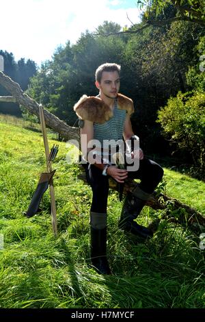 Mittelalterliche jungen ein Mann gekleidet, Trinkhorn hoch in der Hand hält und sitzt auf Ast in der Natur Stockfoto