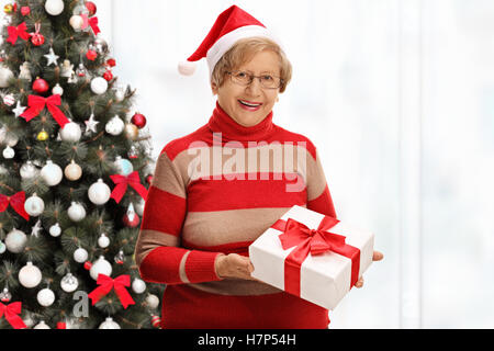 Freudige Seniorin hält ein Weihnachtsgeschenk vor einem Weihnachtsbaum Stockfoto