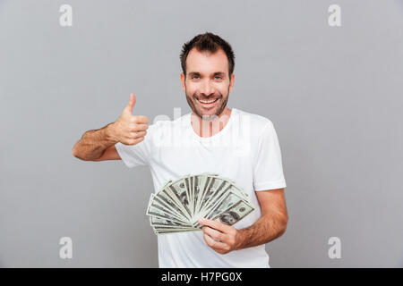 Fröhlicher erfolgreichen junger Mann halten Geld und zeigt Daumen hoch über grauen Hintergrund Stockfoto