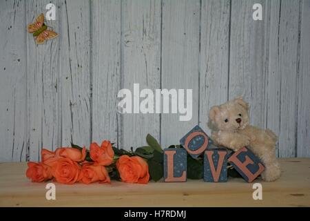 Weißer Teddybär basiert auf Cube mit dem Wort Liebe vor weißen Holzwand mit orange Rosen Stockfoto