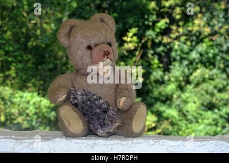 Teddy Bär sitzt auf einer weißen Wand vor grünen Hintergrund und Amethyst in der hand hält Stockfoto