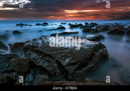 Wasser umgibt die Felsen am Strand, und die Wolken leuchten Orange und rosa bei Sonnenuntergang über dem Horizont; Tarifa, Cádiz, Andalusien, Spanien Stockfoto