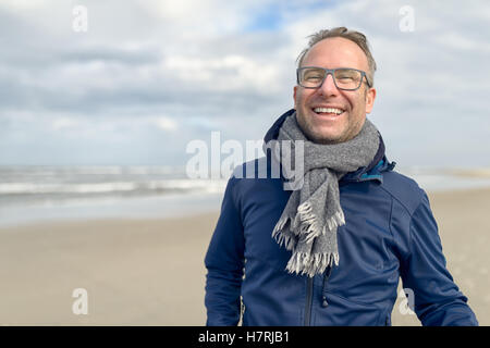 Glücklich lachende Mann mittleren Alters mit Brille und einem gestrickten wollenen Schal stehend auf einem verlassenen Herbst Strand an einem bewölkten Tag w Stockfoto