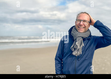Glücklich lachende Mann mittleren Alters mit Brille und einem gestrickten wollenen Schal stehend auf einem verlassenen Herbst Strand an einem bewölkten Tag w Stockfoto