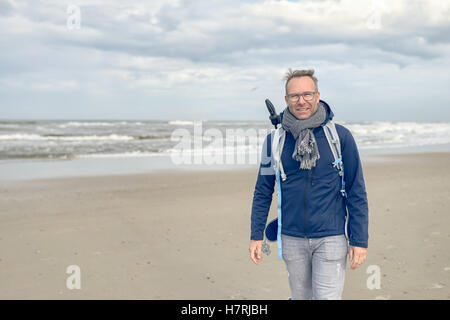 Im mittleren Alter Mann mit Brille, einen Rucksack und einen Schal zu Fuß entlang eines kalten blustery Strandes an einem regnerischen trüben Herbst oder winter Stockfoto