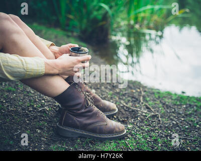Eine junge Frau sitzt an einem Teich im Wald und ist mit einer Tasse Tee Stockfoto