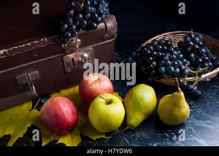 Reife Früchte, Äpfel, Birnen und mehrere gelbe Herbst Blätter auf dunklem Marmor. Reife Trauben in gelben Korb Stockfoto