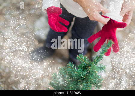 Fürsorgliche Mutter Kind in der Nähe von kleinen Weihnachtsbaum abstrakt mit Schnee-Effekt rote Handschuhe anziehen. Stockfoto