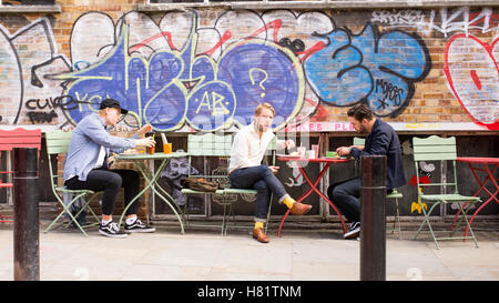 Junge Männer, die einen Brunch, draußen zu sitzen, vor einer Wand bedeckt Graffiti im kühlen Bereich der Shoreditch, East London Stockfoto