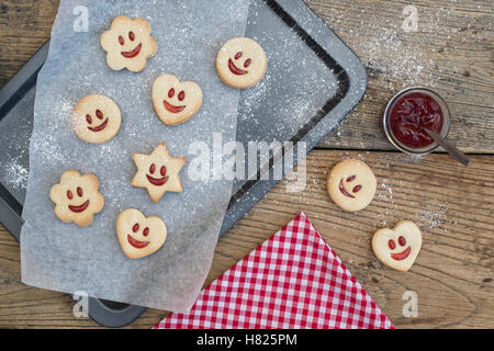 Hausgemachte Jammie Dodgers. Lächelndes Gesicht Kekse und Puderzucker mit einem Topf Marmelade auf Holz Stockfoto