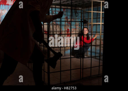 Schattenhafte männliche Figur mit einer Axt vor Angst weibliche Opfer eingesperrt in einen Metall-Käfig mit Blut bespritzt Wand hinter ihr sitzen in Terror ein Schicksal erwartet. Stockfoto