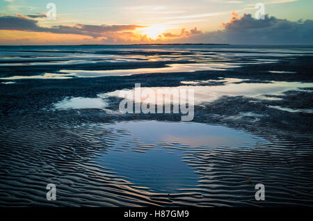 Flinthorndünen, Langeoog. Deutschland. Deutschland. Ein Blick von der Buche bei Ebbe mit dem Sonnenuntergang über der benachbarten Insel Baltrum. Die warme Sonne auf dem Wasser spiegelt, während das Meer rund um die blaufärbung Himmel spiegelt. Tiefe Muster in der Sandstrand lassen flache Becken mit Wasser, die auch sind voll von Reflexionen. Stockfoto