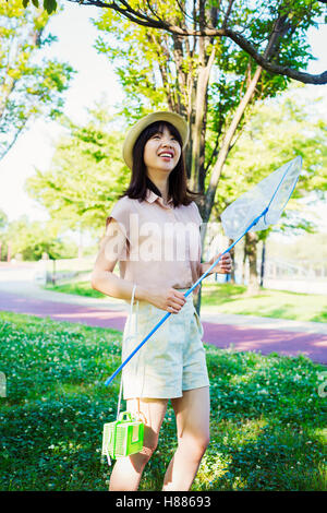 Junge Frau mit langen braunen Haaren steht in einem Park, hält ein Schmetterlingsnetz. Stockfoto