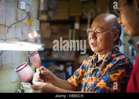 Zwei Personen, Vater und Sohn bei der Arbeit in einem Glasmacher Studio Workshop Inspektion ein rotes geschliffenes Glas Wein Glas. Stockfoto