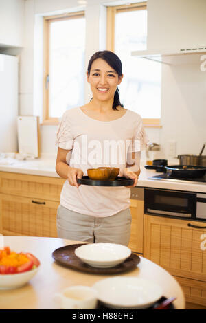 Haus der Familie. Eine Frau, die Zubereitung einer Mahlzeit arrangieren Gerichte auf den Tisch. hält ein schwarzes rundes Tablett Stockfoto