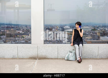 Eine Frau sitzt an einem Fenster mit Blick über eine große Stadt mit dem Rücken zur Ansicht, tätigen eines Anrufs auf einem Smartphone. Stockfoto
