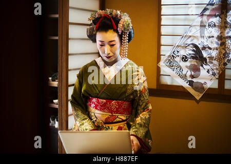 Eine Frau gekleidet im Stil traditionellen Geisha trägt einen Kimono und Obi mit einem aufwendigen Frisur und blumige Haarspangen mit weißem Gesicht Make-up mit knallroten Lippen und dunkle Augen mit einem Laptopcomputer. Stockfoto