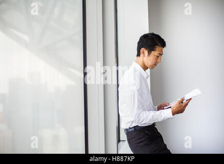 Ein Geschäftsmann im Büro durch ein großes Fenster lesen Papierkram. der Fensterbank gelehnt. Stadtbüro. Arbeitsleben Stockfoto