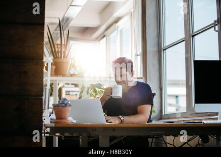 Entspannte junger Mann sitzt an seinem Schreibtisch arbeiten am Laptop und Kaffee trinken. Geschäftsmann in modernen Arbeitsplatz. Stockfoto