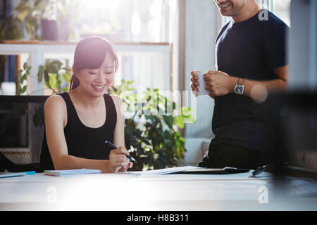 Schuss der lächelnde junge Frau sitzt an ihrem Schreibtisch arbeiten mit männlichen Kollegen stehen. Geschäftsleute bei der Arbeit im Büro. Stockfoto