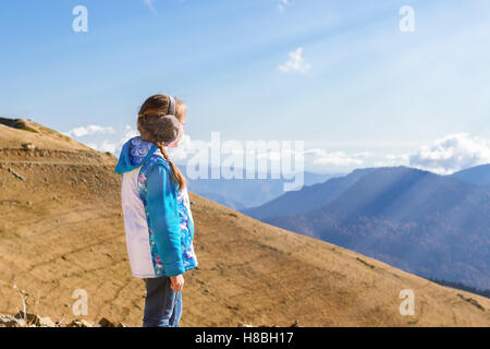 Mädchen mit einem Zopf, gekleidet in Sportjacke und warmen flauschigen Kopfhörer bewundert herbstliche Berglandschaft, Sotschi, Russland Stockfoto